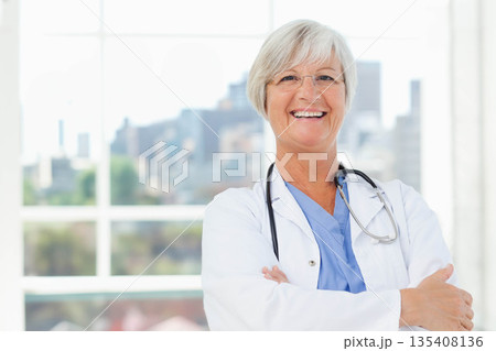 Senior female doctor standing wearing lab coat and blue scrubs by window with stethoscope smiling 135408136