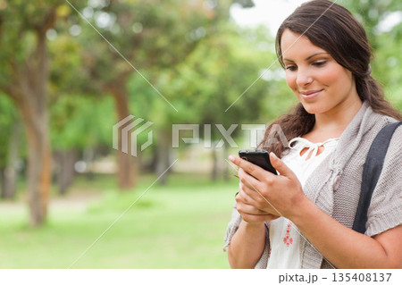 Woman holding smartphone while looking at screen in grassy park with backpack strap, copy space Woman holding smartphone while looking at screen in grassy park with backpack strap, copy space 135408137