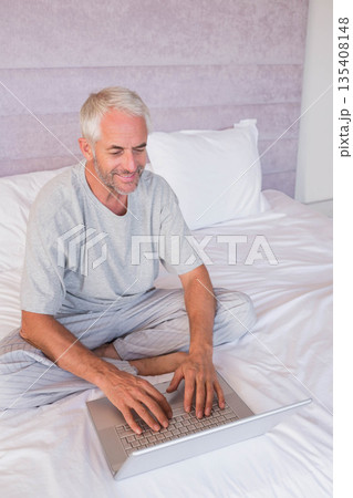 Senior man in pajamas typing on laptop in bedroom while sitting cross-legged on bed with pillows Senior man in pajamas typing on laptop in bedroom while sitting cross-legged on bed with pillows 135408148