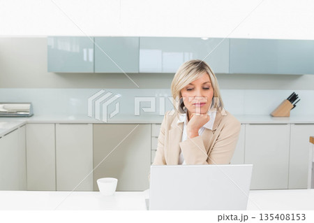 Woman working on laptop at modern home kitchen table with white mug and wooden knife block 135408153