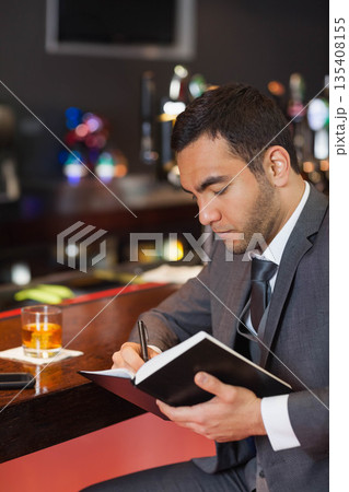 Middle Eastern man in gray suit sitting at wooden bar counter writing in notebook with drink Middle Eastern man in gray suit sitting at wooden bar counter writing in notebook with drink 135408155