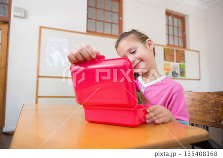Female child student opening red plastic lunchbox while sitting at wooden school desk in classroom 135408168