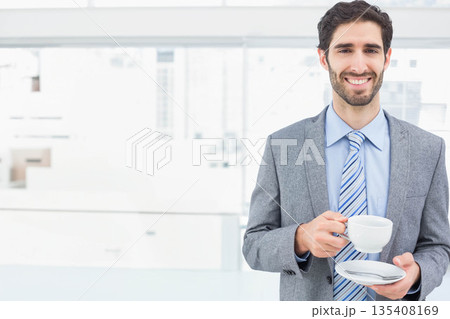 Businessman holding white coffee cup and saucer in office by glass windows wearing suit, copy space Businessman holding white coffee cup and saucer in office by glass windows wearing suit, copy space 135408169