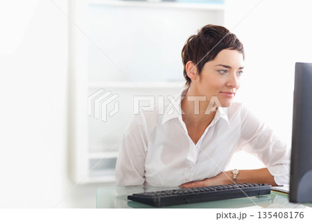 Mid-adult white woman checking watch and typing on computer at glass office desk, copy space Mid-adult white woman checking watch and typing on computer at glass office desk, copy space 135408176