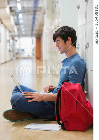 Male student sitting in hallway studying on laptop next to red backpack and papers, copy space 135408181