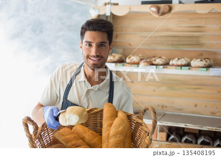 Male baker holding baguette basket wearing apron and blue glove in bakery by shelves with loaves 135408195