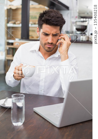 Businessman using smartphone for phone call while sipping coffee at coffee shop table with laptop 135408218