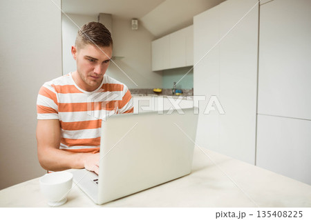 Man sitting at kitchen table focusing on silver laptop with mug, fruit bowl and water bottle Man sitting at kitchen table focusing on silver laptop with mug, fruit bowl and water bottle 135408225