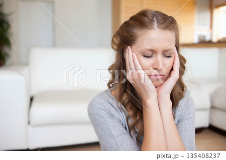 Woman wearing grey top pressing hands against cheeks on floor in living room before white sofa 135408237