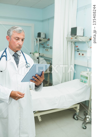 Senior male doctor wearing lab coat checking tablet at hospital bedside with IV pump, copy space Senior male doctor wearing lab coat checking tablet at hospital bedside with IV pump, copy space 135408244
