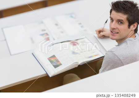 Male student writing notes with pen looking toward camera at lecture desk with textbook, copy space Male student writing notes with pen looking toward camera at lecture desk with textbook, copy space 135408249