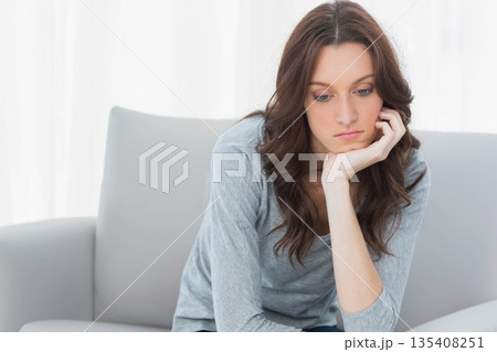Woman leaning forward resting chin on hand on sofa in bright living room with sheer curtains 135408251