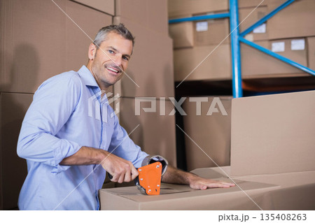 Middle-aged man using orange tape dispenser sealing cardboard boxes at warehouse with blue shelving Middle-aged man using orange tape dispenser sealing cardboard boxes at warehouse with blue shelving 135408263