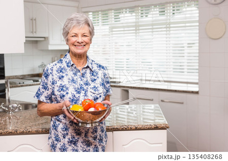 Senior woman holding steel colander filled with red tomatoes and yellow bell pepper in home kitchen Senior woman holding steel colander filled with red tomatoes and yellow bell pepper in home kitchen 135408268