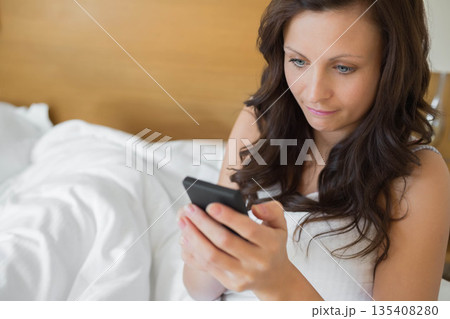 Woman sitting on bed holding smartphone and reading messages in bedroom, copy space 135408280