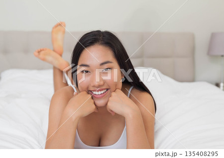 Asian woman lying on bed in bedroom propping chin on fists with white duvet, lavender lamp 135408295