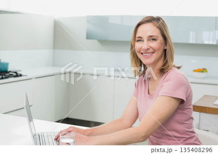 Woman smiling at camera while working on laptop at kitchen counter with fruit bowl, copy space 135408296