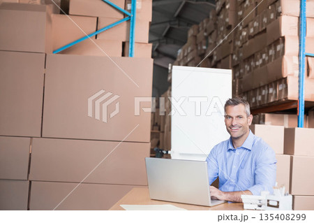 Middle-aged man sitting at desk in warehouse using laptop with whiteboard behind, copy space 135408299