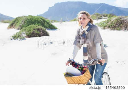 Mature white woman riding bicycle on dune with wicker basket holding blanket and wide-brimmed hat Mature white woman riding bicycle on dune with wicker basket holding blanket and wide-brimmed hat 135408300