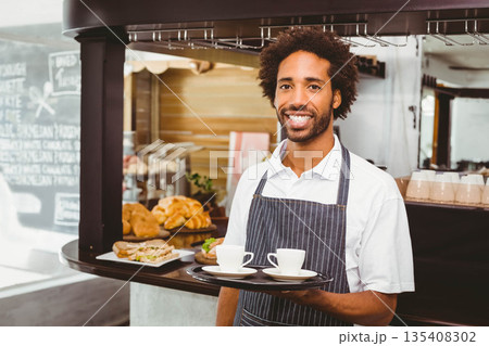 African American man wearing apron serving white coffee cups on tray at cafe counter with pastries 135408302