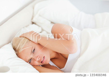Woman lying on bed with white sheets and having headache in bright bedroom with mug beside 135408346