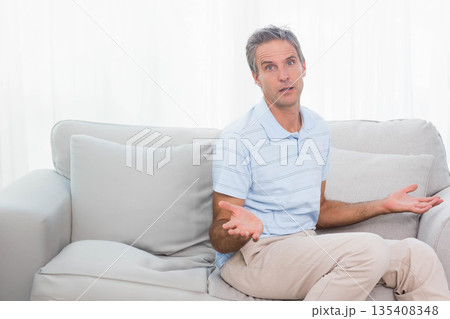 Man raising palms asking question seated on sofa in living room with cushions, curtains, copy space 135408348