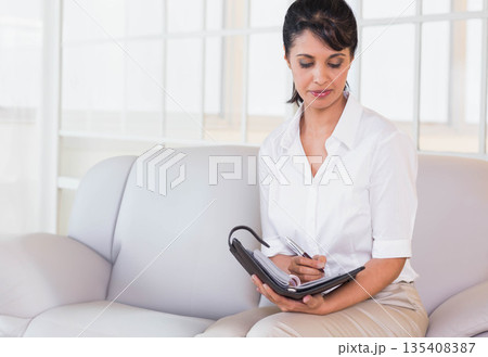 Mid adult woman writing in ring-bound planner with pen on sofa in office lounge, copy space Mid adult woman writing in ring-bound planner with pen on sofa in office lounge, copy space 135408387