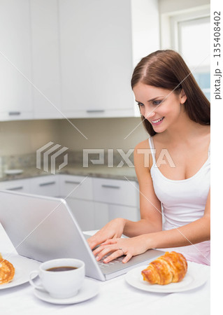 Woman typing on silver laptop at kitchen table with golden croissants and coffee cup, copy space 135408402