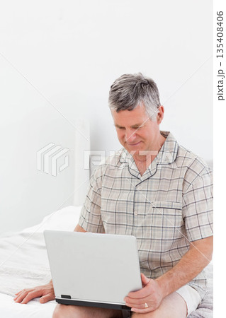 Senior man sitting on bed edge using laptop inside bedroom with crisp white bedding and headboard 135408406