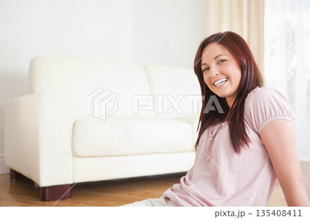 Woman sitting on wooden floor next to white sofa in living room under sheer curtains smiling Woman sitting on wooden floor next to white sofa in living room under sheer curtains smiling 135408411
