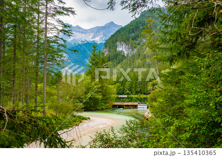 Famous green lake Schiederweiher near village Hinterstoder. Small weir and dam on lake with flowing water. Big Austrian mountains in background. Soft and magical colors in pure nature. 135410365