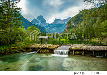 Famous green lake Schiederweiher near village Hinterstoder. Small weir and dam on lake with flowing water. Big Austrian mountains in background. Soft and magical colors in pure nature. Famous green lake Schiederweiher near village Hinterstoder. Small weir and dam on lake with flowing water. Big Austrian mountains in background. Soft and magical colors in pure nature. 135410368