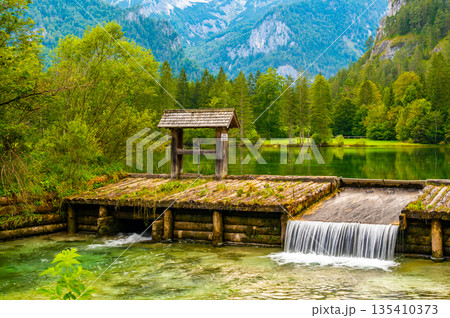 Famous green lake Schiederweiher near village Hinterstoder. Small weir and dam on lake with flowing water. Big Austrian mountains in background. Soft and magical colors in pure nature. Famous green lake Schiederweiher near village Hinterstoder. Small weir and dam on lake with flowing water. Big Austrian mountains in background. Soft and magical colors in pure nature. 135410373