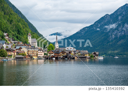 Hallstatt Village and Hallstatter See lake in Austria. Scenery with famous old church near the lake. Clouds and mist over the mountains in background. Famous tourist destination. Hallstatt Village and Hallstatter See lake in Austria. Scenery with famous old church near the lake. Clouds and mist over the mountains in background. Famous tourist destination. 135410392