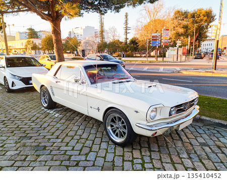 Elegant white classic muscle car parked on urban cobblestones under a warm sunset glow. Symbolizing freedom and retro luxury, this image is ideal for lifestyle and automotive marketing. Elegant white classic muscle car parked on urban cobblestones under a warm sunset glow. Symbolizing freedom and retro luxury, this image is ideal for lifestyle and automotive marketing. 135410452
