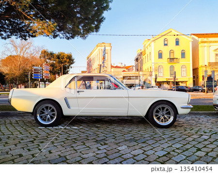 Stunning profile of a classic white muscle car parked on a European cobblestone street at sunset. This nostalgic image evokes feelings of freedom, vintage style, and urban elegance. 135410454