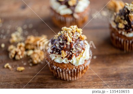 Tasty chocolate muffin with creme mascarpone and chocolate on top, covered with nuts. Placed on wooden desk. Sweet fresh and homemade cupcake. 135410655