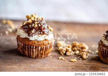 Tasty chocolate muffin with creme mascarpone and chocolate on top, covered with nuts. Placed on wooden desk. Sweet fresh and homemade cupcake. 135410661