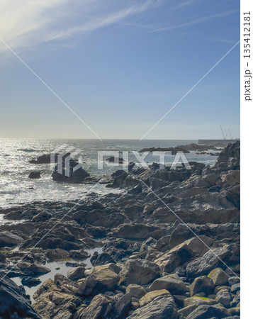 Rocky shoreline with rugged boulders stretching into the sea, waves shimmering under the bright Rocky shoreline with rugged boulders stretching into the sea, waves shimmering under the bright 135412181