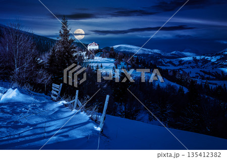 countryside in mountains at night. lovely scenery with wooden fence and coniferous forest on a snowy slopes in full moon light. village on distant hill. finance investment or abstract concepts countryside in mountains at night. lovely scenery with wooden fence and coniferous forest on a snowy slopes in full moon light. village on distant hill. finance investment or abstract concepts 135412382