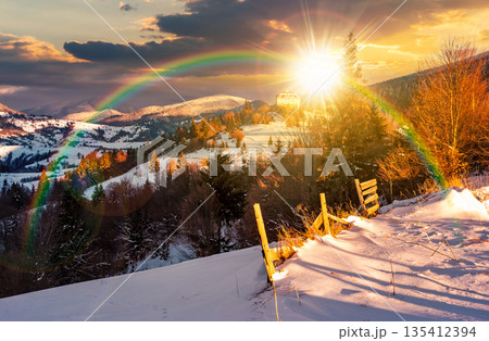 countryside in mountains at sunset. lovely scenery with wooden fence and coniferous forest on a snowy slopes in evening light. village on distant hill. leadership corporate wallpaper under the rainbow 135412394