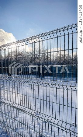 Metal wire fence covered in thick transparent ice glaze against a snowy park and sunny winter sky Metal wire fence covered in thick transparent ice glaze against a snowy park and sunny winter sky 135413459