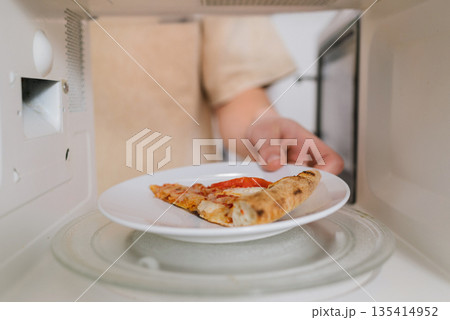 Man placing a white plate with a slice of pizza into a microwave for quick reheating. Man placing a white plate with a slice of pizza into a microwave for quick reheating. 135414952