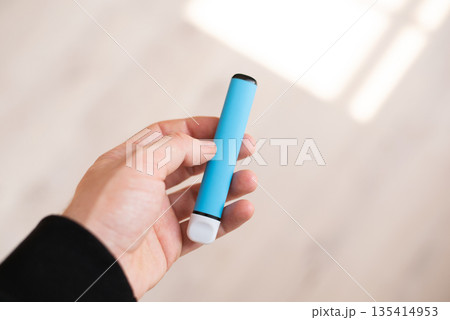 Close-up of a hand holding a blue disposable electronic cigarette or vape pen. Close-up of a hand holding a blue disposable electronic cigarette or vape pen. 135414953