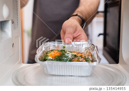 Man placing foil tray with fried eggs into microwave, highlighting unsafe appliance use. Man placing foil tray with fried eggs into microwave, highlighting unsafe appliance use. 135414955