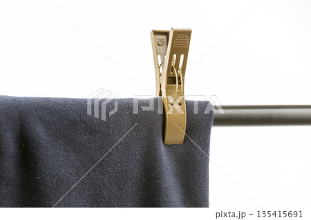 A close-up shows dark blue fabric, possibly garment, secured by a large, tan plastic clothespin on metal rod isolated on a white A close-up shows dark blue fabric, possibly garment, secured by a large, tan plastic clothespin on metal rod isolated on a white 135415691