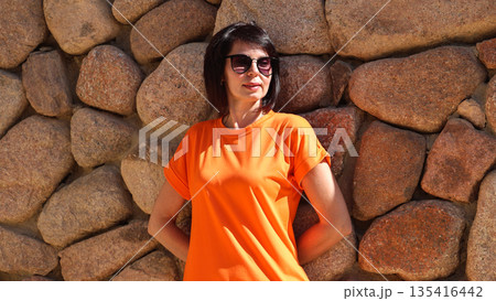 Elegant woman in orange dress standing against rustic stone wall, wearing sunglasses outdoors 135416442