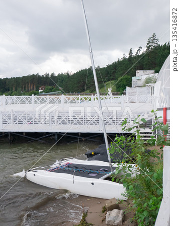 White catamaran sailboat on river shore with water lapping at the hull. Outdoor recreation and water sport concept for travel brochure. White catamaran sailboat on river shore with water lapping at the hull. Outdoor recreation and water sport concept for travel brochure. 135416872