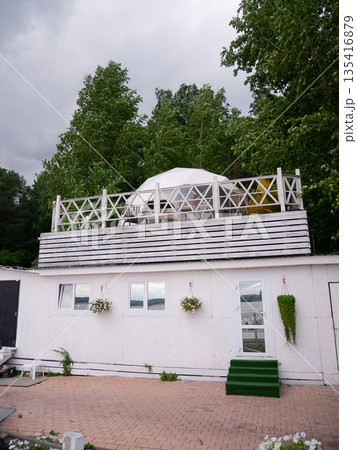 White modular house with outdoor terrace and geodesic dome on the roof, surrounded by green trees, under a cloudy sky. Summer vacation rental home. White modular house with outdoor terrace and geodesic dome on the roof, surrounded by green trees, under a cloudy sky. Summer vacation rental home. 135416879