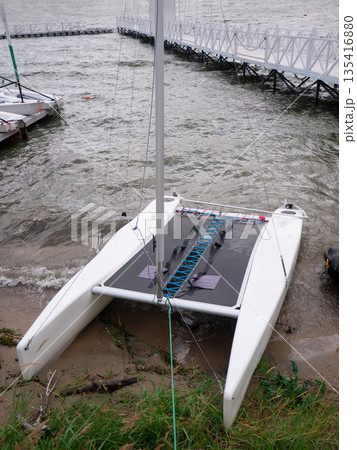 Small sailing catamaran docked on sandy shore next to choppy water. Recreational watersport boat for hobby and leisure activity. Small sailing catamaran docked on sandy shore next to choppy water. Recreational watersport boat for hobby and leisure activity. 135416880